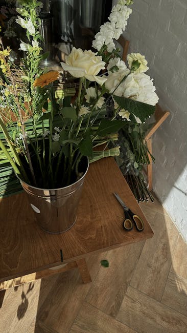 A tall, elegant bouquet arrangement placed in a shiny, metallic vase sits on a wooden table. The bouquet features a mix of large, creamy white roses and delicate white lisianthus, complemented by tall