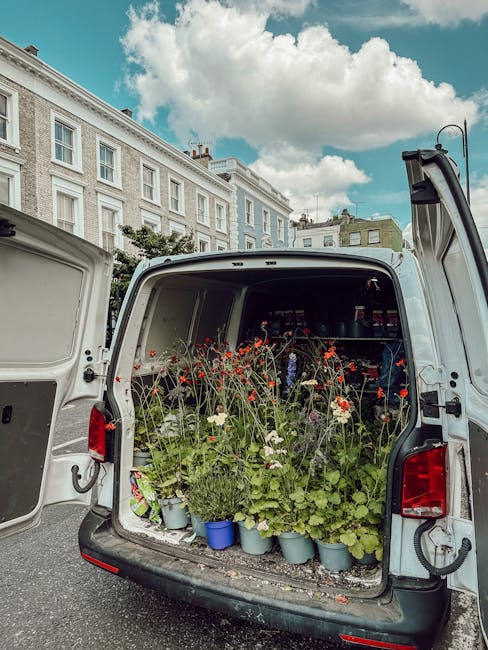 The open rear of a white delivery van filled with potted flowers, including red poppies, white daisies, and green foliage, arranged in blue and terracotta pots. The flowers are fresh, with vibrant pet