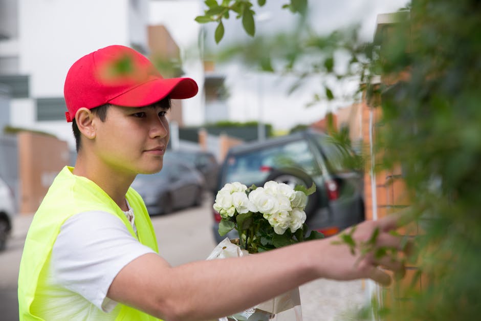 A young male flower delivery worker wearing a red cap and a high-visibility yellow vest is seen outdoors, carefully placing a bouquet of white hydrangeas against a flowering shrub. The bouquet feature