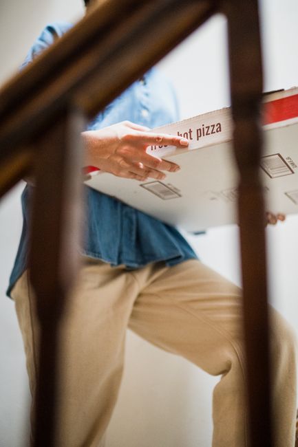 A person wearing a light blue denim shirt and beige trousers is holding a white pizza box with red text, positioned on their left side, with their hand gripping the side. The scene is viewed through a
