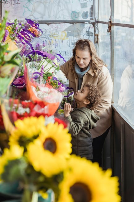A woman and a young girl stand together inside a flower shop, surrounded by vibrant bouquets of fresh flowers including sunflowers, purple irises, red roses, and mixed seasonal blooms. The flowers are
