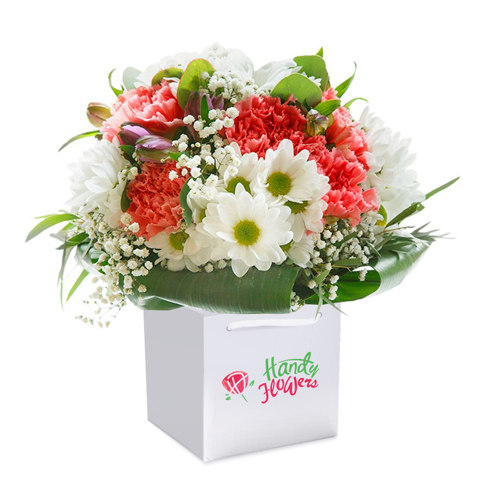 Beautifully styled and meticulously detailed, this bouquet delivers the natural beauty and cheerful charm representative of Bishop's Stortford floristry. Presented upright in a fresh, square white gift bag equipped with soft ribbon handles, the bouquet's heart lies in an artful blend of coral and peach carnations, each bloom unfurling velvety, frilled petals that emanate softness and lush texture. Bright white daisies with eye-catching lime-green centers dot the canvas, creating a sharp and uplifting contrast that reflects the cheerful vibe of the nearby Jackson Square markets. Airy white baby's breath intertwines effortlessly through the bouquet, offering a weightless, cloud-like quality reminiscent of a gentle, misty morning at Castle Park. Nestled among the main flowers on the left, several unopened, purple-tinged buds add gentle intrigue and echo the quiet promise found in a garden on the edge of spring. Broad, glossy deep green foliage frames the base, supporting and accentuating the freshness of each bloom. The arrangement stands as a symbol of skilled floristry and attention to detail-its balanced, rounded composition ideal for marking personal celebrations such as birthdays and anniversaries or bringing everyday delight to households across Bishop's Stortford. The luminous lighting enhances every shade and shape, providing clarity that evokes the burst of life seen in the town's best flower displays. Suitable for seamless local delivery or a heartfelt handoff, this splendid bouquet imaginatively captures the town's vibrant spirit.