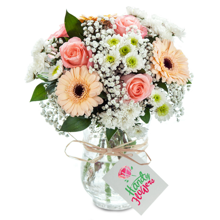 Positioned against a flawless white background, this expertly arranged bouquet seems to capture the very essence of Bishop's Stortford's gentle beauty. The centerpiece is a cluster of classic, peach-toned roses-softly blushing and romantically open, their petals like silk in the morning light. Two prominent gerbera daisies, each hued in warm peach with chocolate-brown hearts, draw the eye with cheerful simplicity, echoing the neighborly vibrancy of Jackson Square. Intermingled among these are generous clouds of white baby's breath, airy and light, adding fullness and a whisper of nostalgia to the ensemble. A scattering of petite white chrysanthemums, their lemon-lime centers peering out with fresh optimism, lends textural depth and life, reminding one of spring blooms in Sworders Field. Carefully placed among the flowers, lush green eucalyptus leaves impart a rustic woodland freshness, their scent subtle and comforting. The stems, visible through a gently patterned glass vase, tell the story of loving craftsmanship and daily dedication typical of your area's family-run florists. At the neck, a neat bow of pale raffia holds a simple white gift tag, inviting a personal message-perhaps for a birthday, a thank-you, or to mark a local anniversary. This arrangement, equally suited for intimate home gatherings or thoughtful deliveries throughout Bishop's Stortford, celebrates both the artistry and heart of local floristry.